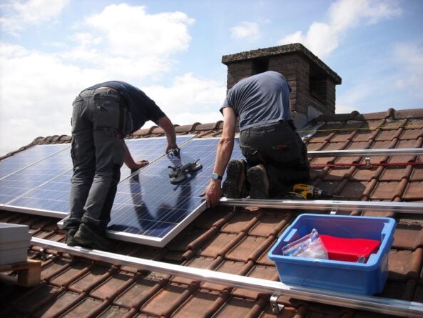Two people installing solar PV panels on a roof with several pieces of equipment.
